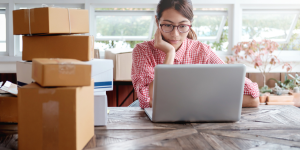A woman sitting looking at her laptop with packages sat on her desk beside her